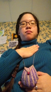 a brunette girl with glasses and long hair in a braid wearing a turquoise turtleneck and a purple beaded necklace purse has one hand on their upper chest and one hand on their stomach